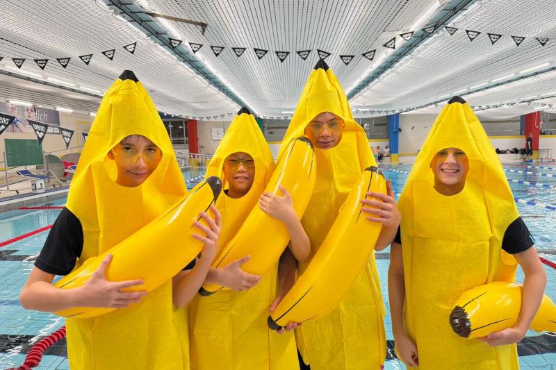 Vier Personen in Bananenkostümen stehen am Beckenrand eines Schwimmbads und halten gelbe Bananen in den Händen.