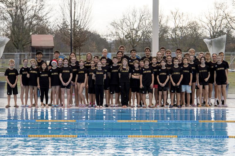 Gruppenfoto von Schwimmern in schwarzen T-Shirts am Poolrand, hinter ihnen ein Schwimmbecken und Bäume im Hintergrund.