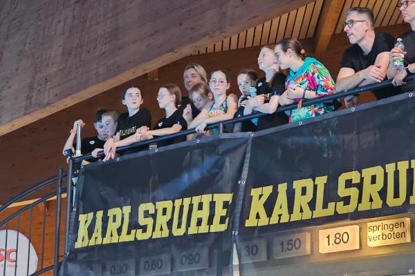 Junge Zuschauer stehen am Balkon einer Sporthalle mit einem Banner 'Karlsruhe'. Treppen im Hintergrund sichtbar.