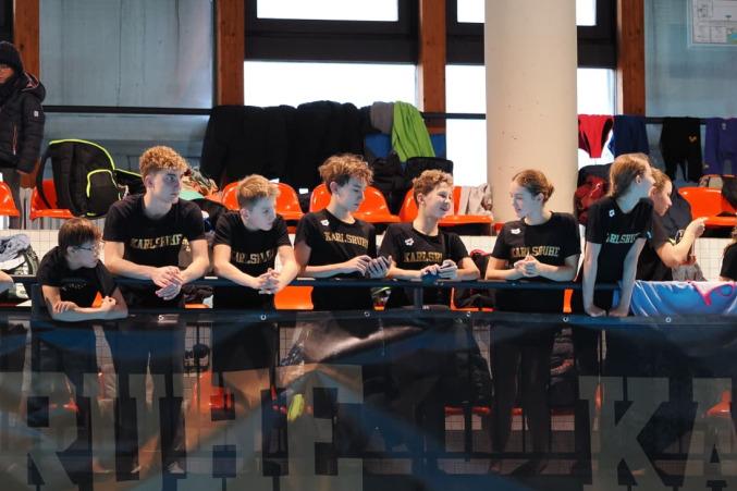 Gruppe junger Sportler in schwarz-gelben T-Shirts lehnt an einer Balustrade in einer Sporthalle.