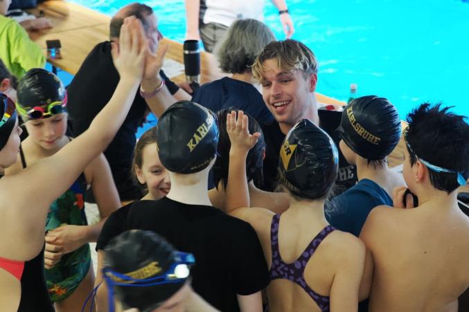 Gruppe von jungen Schwimmern mit Badekappen feiert an einer Schwimmbeckenrand, lächeln und klatschen in die Hände.