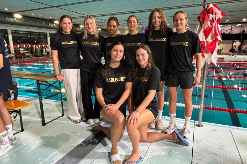 Gruppensfoto von acht Schwimmerinnen in schwarzen T-Shirts mit „Karlsruhe“ am Beckenrand eines Schwimmbads.