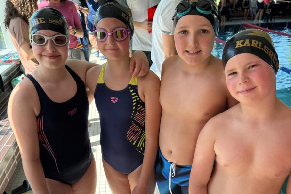 Vier Kinder in Schwimmbekleidung und Badekappen stehen am Rand eines Schwimmbeckens. Zuschauer sind im Hintergrund sichtbar.