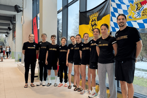 Gruppe junger Sportler in schwarzen T-Shirts mit "Karlsruhe" vor Fenstern und Fahnen in einem Sportzentrum.