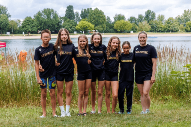 Gruppe junger Sportler in schwarzen T-Shirts mit &bdquo;Karlsruhe&ldquo; am Wasser, umgeben von gr&uuml;ner Landschaft.