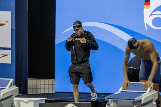 Ein Schwimmer in schwarzer Trainingskleidung betritt die Wettkampfb&uuml;hne mit einem Handtuch in der Hand.