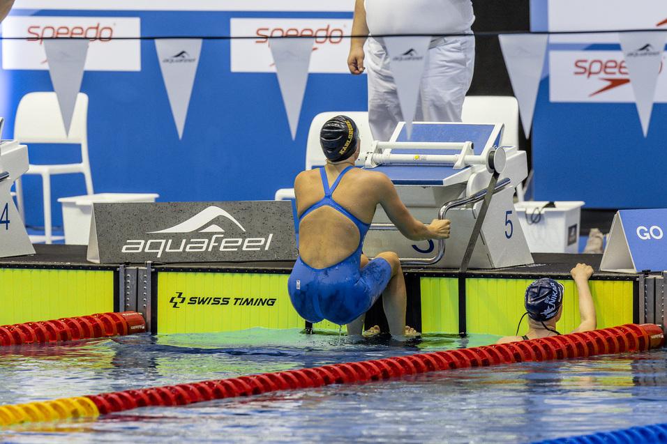 Schwimmerin in blauer Badebekleidung sitzt am Beckenrand, bereit zum Start, w&auml;hrend ein Coach sie beobachtet.