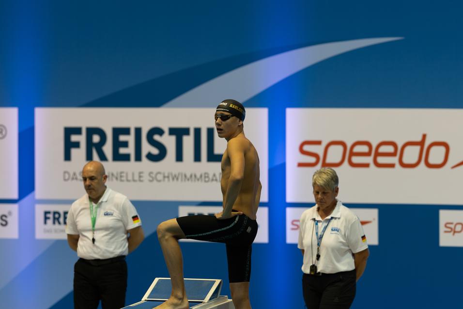 Junger Schwimmer steht auf dem Startblock, bereit zum Sprung, w&auml;hrend zwei Trainer ihn anfeuern.