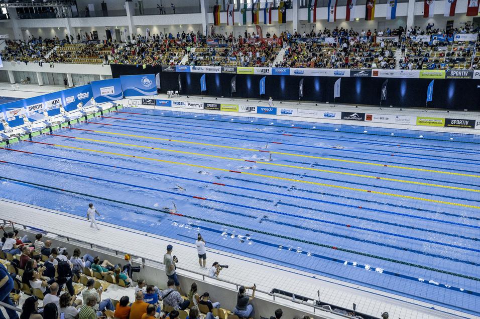 Schwimmbecken mit mehreren Bahnen, besetzt mit Schwimmern, und Publikum auf Trib&uuml;nen in einer Sportarena.