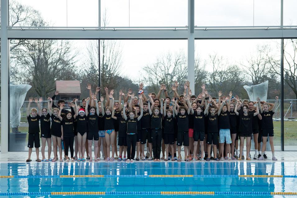 Gruppe von Schwimmern in schwarzen T-Shirts, die am Beckenrand jubeln, mit Glasw&auml;nden und Au&szlig;enblick im Hintergrund.