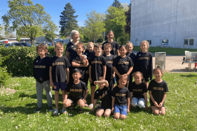 Gruppe von Kindern in schwarzen T-Shirts mit dem Schriftzug "Karlsruhe", posierend auf einer Wiese an einem sonnigen Tag.