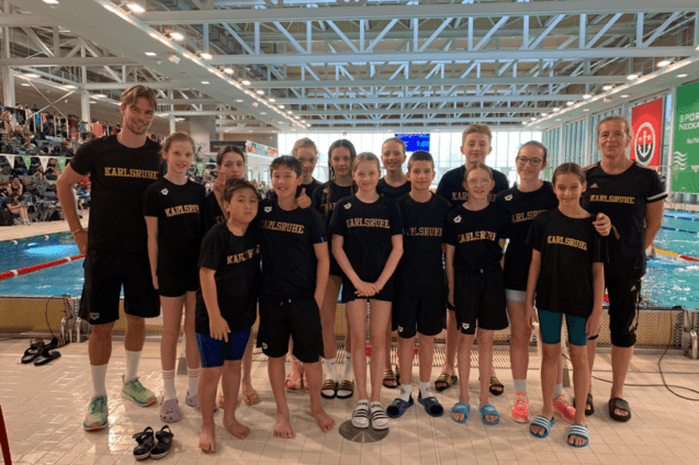 Gruppe von Kindern und Trainern in schwarzen T-Shirts mit der Aufschrift &bdquo;Karlsruhe&ldquo; am Schwimmbecken.