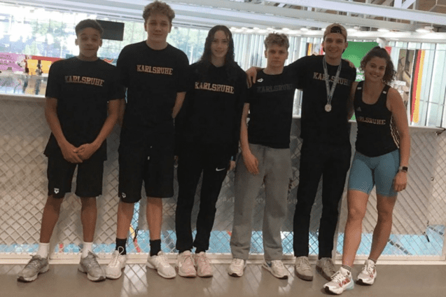 Sechs Jugendliche mit Sportbekleidung stehen l&auml;chelnd vor einer Schwimmhalle, einige tragen Medaillen.