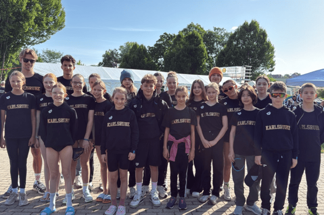 Gruppenfoto von Jugendlichen in schwarzen Trainingsanz&uuml;gen mit &bdquo;Karlsruhe&ldquo;-Aufdruck, stehend auf einem Sportgel&auml;nde.