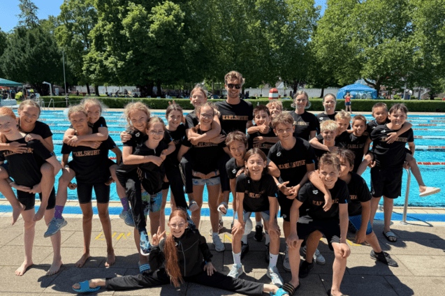 Eine Gruppe von Kindern und Jugendlichen in schwarzen T-Shirts mit &bdquo;Karlsruhe&ldquo; posiert l&auml;chelnd am Schwimmbecken.