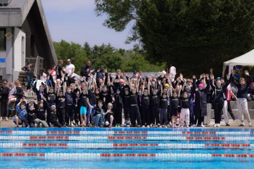 Eine Gruppe von Kindern und Betreuern steht am Schwimmbecken, einige halten Medaillen und alle heben die H&auml;nde.