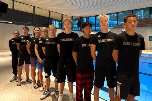 Gruppierung von zehn jungen Sportlern in schwarzen T-Shirts mit "Karlsruhe" am Beckenrand eines Schwimmbads.