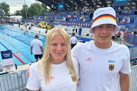 Zwei jugendliche Schwimmer posieren am Beckenrand eines Schwimmwettbewerbs, beide tragen wei&szlig;e T-Shirts und tragen sportliche Kleidung.