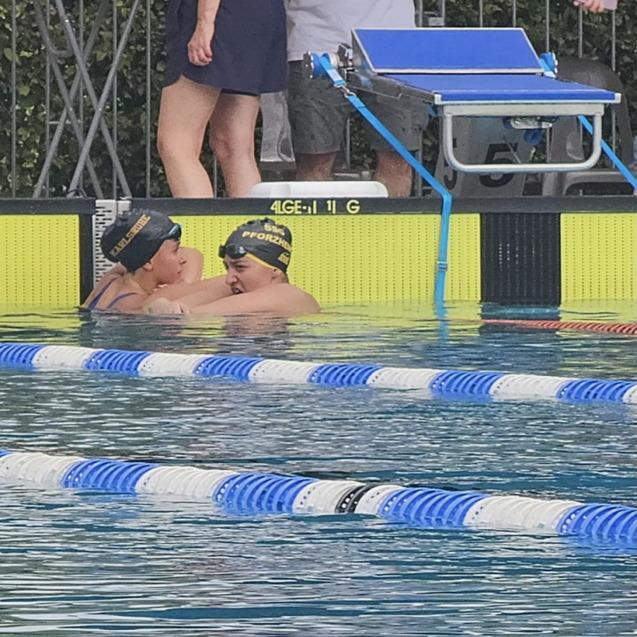 Zwei Schwimmerinnen in Badekappen umarmen sich im Wasser eines Schwimmbeckens, im Hintergrund stehen Zuschauer.