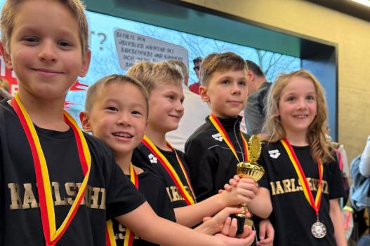 Gruppe von f&uuml;nf Kindern in Sportkleidung mit Medaillen und einem Pokal in der Hand, l&auml;chelnd vor einem Hintergrund.