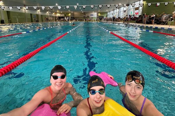 Drei schwimmende Kinder mit Schwimmbrettern posieren am Beckenrand eines Hallenbades.