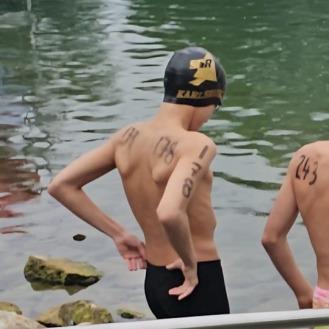 Zwei Schwimmer stehen am Ufer eines Gew&auml;ssers, einer tr&auml;gt eine Badekappe und hat Wettkampfnumbers auf den Armen.