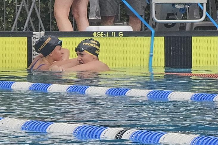 Zwei Schwimmerinnen in Badekappen umarmen sich im Wasser eines Schwimmbeckens, im Hintergrund stehen Zuschauer.