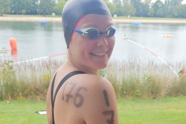 Wettkampfschwimmerin in schwarzem Anzug mit Schwimmhaube, Zahlen auf dem Arm, steht am Ufer eines Gew&auml;ssers.