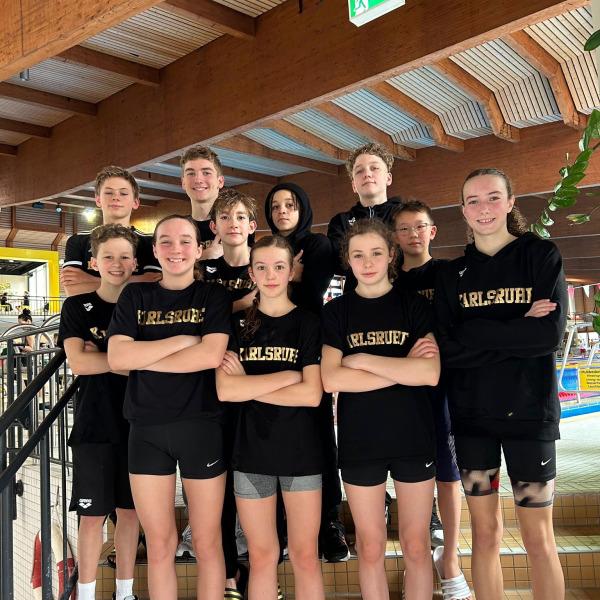 Gruppe von zehn Jugendlichen in schwarzen T-Shirts mit Aufdruck „Karlsruhe“ posiert auf einer Treppe in einem Schwimmbad.