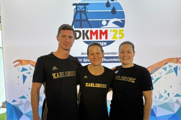 Drei Schwimmer in schwarzen Karlsruher Trikots stehen vor einem Banner der DKMM '25 im Schwimmsportzentrum.