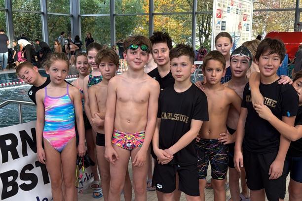 Gruppe von Kindern in Schwimmbekleidung, l&auml;chelnd vor einem Schwimmbecken und einem Sponsorenbanner.