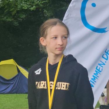 M&auml;dchen in schwarzem Hoodie mit Medaille steht vor Zelt und Banner im Freien.