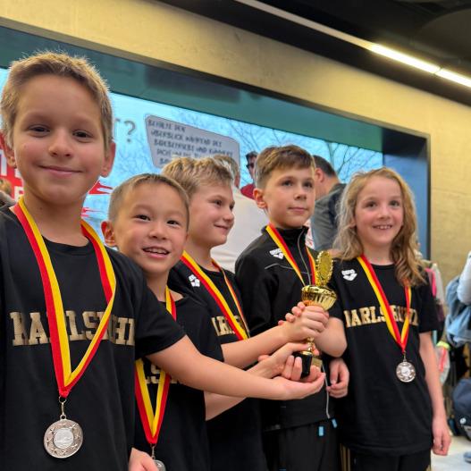 Gruppe von f&uuml;nf Kindern in Sportkleidung mit Medaillen und einem Pokal in der Hand, l&auml;chelnd vor einem Hintergrund.
