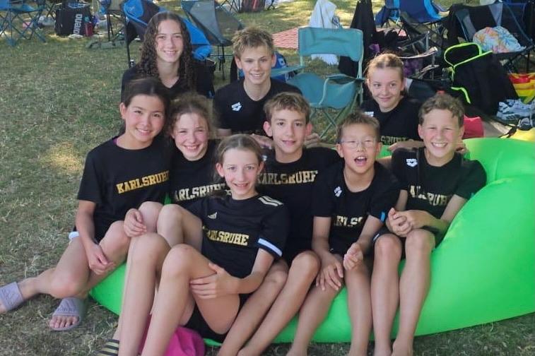 Gruppe von neun Kindern in T-Shirts mit Aufdruck &bdquo;Karlsruhe&ldquo; auf einer Wiese, umgeben von Campingst&uuml;hlen.