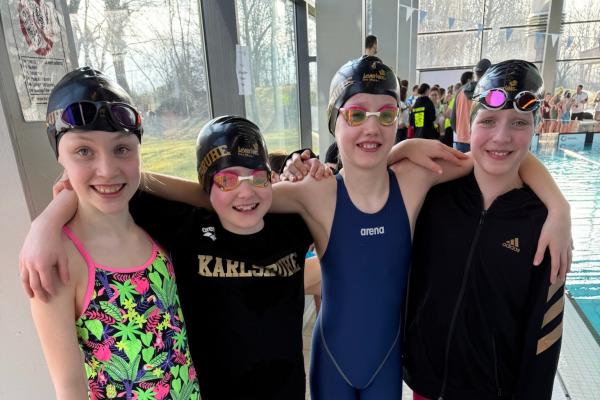 Vier Schwimmerinnen in Schwimmbekleidung mit Badekappen stehen l&auml;chelnd am Rand eines Schwimmbeckens.