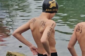 Zwei Schwimmer stehen am Ufer eines Gew&auml;ssers, einer tr&auml;gt eine Badekappe und hat Wettkampfnumbers auf den Armen.