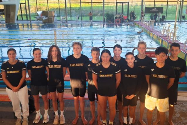 Gruppe von zw&ouml;lf Schwimmern in schwarzen T-Shirts mit &bdquo;Karlsruhe&ldquo; vor einem Wettkampfbecken.