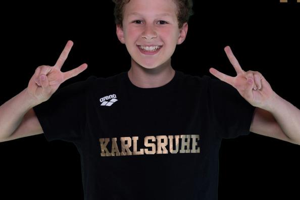 Junge mit lockigem Haar macht Peace-Zeichen, tr&auml;gt schwarzes T-Shirt mit "Karlsruhe" und Hintergrund in Schwarz.