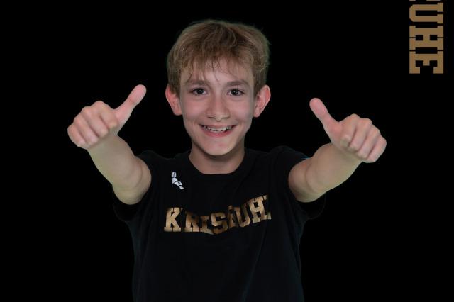 Junge mit Daumen hoch, tr&auml;gt schwarzes Shirt mit Schriftzug &bdquo;Karlsruhe&ldquo;, Hintergrund mit Text zu Schwimmmeisterschaften.
