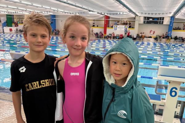 Drei Kinder stehen am Schwimmbecken: ein Junge in schwarzem Shirt, ein M&auml;dchen in pinkem Badeanzug und ein Junge in gr&uuml;nem Poncho.