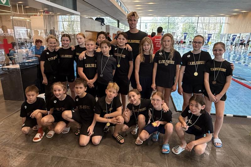 Gruppe von Schwimmern in schwarzen T-Shirts mit Aufschrift &bdquo;Karlsruhe&ldquo; und Medaillen in einem Schwimmbad.