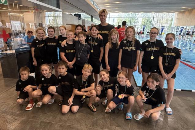Gruppe von Schwimmern in schwarzen T-Shirts mit Aufschrift &bdquo;Karlsruhe&ldquo; und Medaillen in einem Schwimmbad.