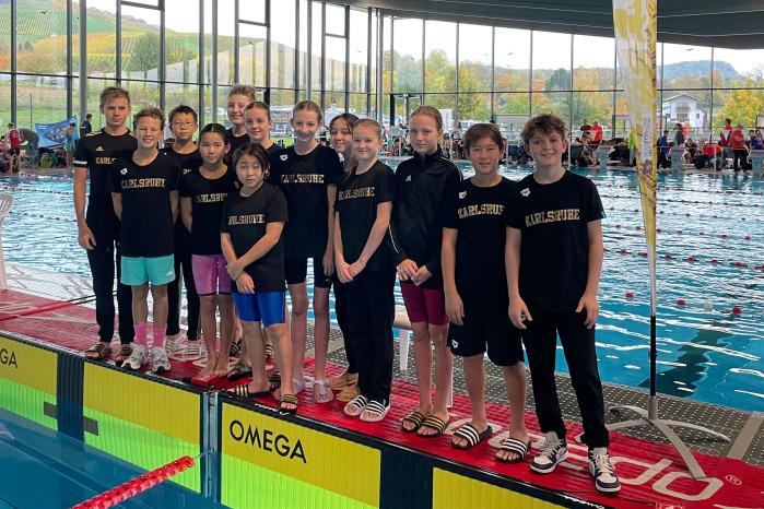 Gruppe junger Schwimmer in einheitlichen T-Shirts mit &bdquo;Karlsruhe&ldquo; stehen am Beckenrand in einem modernen Schwimmbad.