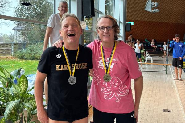 Zwei lächelnde Schwimmer mit Medaillen stehen in einem Schwimmbad, im Hintergrund weitere Teilnehmer und große Fenster.