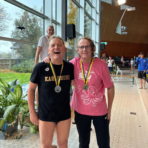 Zwei lächelnde Schwimmer mit Medaillen stehen in einem Schwimmbad, im Hintergrund weitere Teilnehmer und große Fenster.