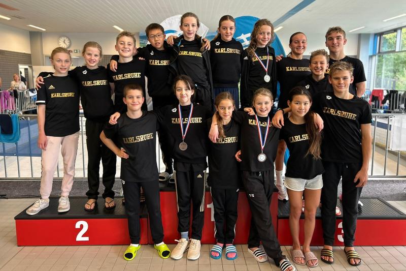 Gruppe von Schwimmsportlern in schwarzen T-Shirts mit der Aufschrift "Karlsruhe" auf dem Siegerpodest nach einem Wettkampf.