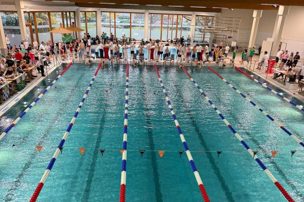 Schwimmbecken mit blauen Wasserbahnen und zahlreichen Zuschauern, die den Wettkampf beobachten. Holzdecke im Hintergrund.