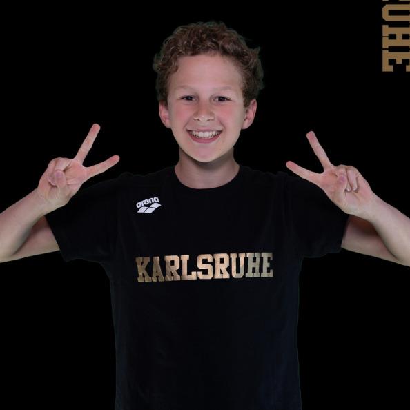 Junge mit lockigem Haar macht Peace-Zeichen, tr&auml;gt schwarzes T-Shirt mit "Karlsruhe" und Hintergrund in Schwarz.