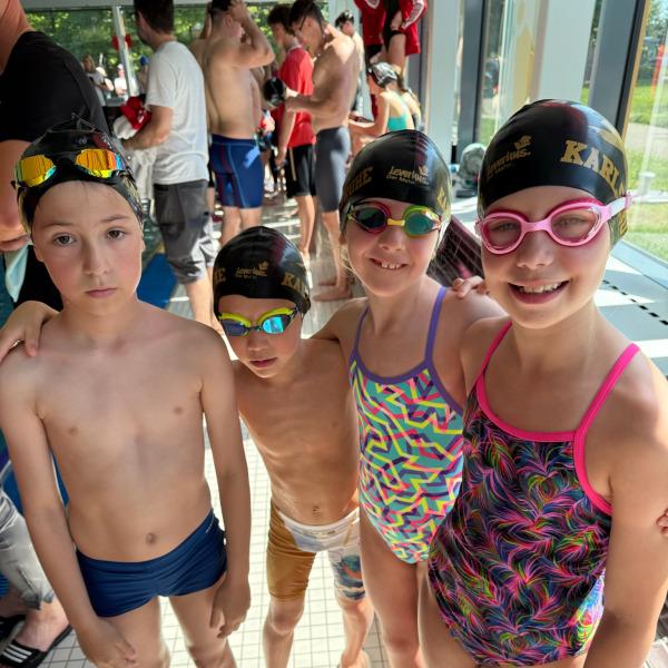 Vier Kinder in Badebekleidung und Schwimmkappen stehen zusammen am Poolrand, fr&ouml;hlich l&auml;chelnd f&uuml;r ein Foto.