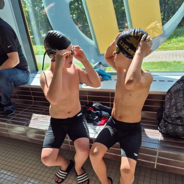 Zwei Jungen sitzen am Beckenrand, setzen Schwimmbrillen auf und tragen Badebekleidung und Badeschuhe.